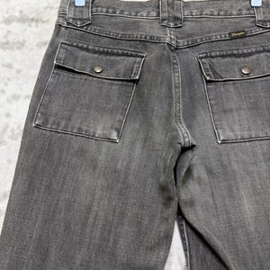 May include: Close-up of the back of gray denim pants with two square pockets and button closures. The Wrangler logo is visible on the back pocket. The pants are hanging on a wooden hanger.