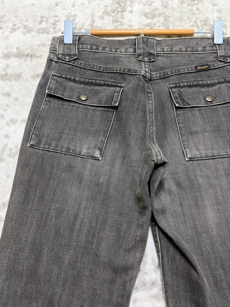 May include: Close-up of the back of gray denim pants with two square pockets and button closures. The Wrangler logo is visible on the back pocket. The pants are hanging on a wooden hanger.