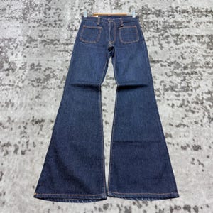 Puede incluir: Vaqueros acampanados de mezclilla azul oscuro con dos bolsillos delanteros y cierre de botón y cremallera. Los vaqueros tienen cintura alta y una amplia campana desde la rodilla. Las costuras son de color naranja en contraste.