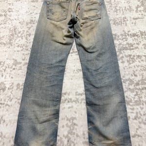 Puede incluir: Vaqueros de mezclilla azul descolorido con detalles desgastados, incluyendo parches desgastados y una etiqueta roja. Los vaqueros se muestran en una percha de madera, mostrando los bolsillos traseros y el estilo vintage. Corte recto.