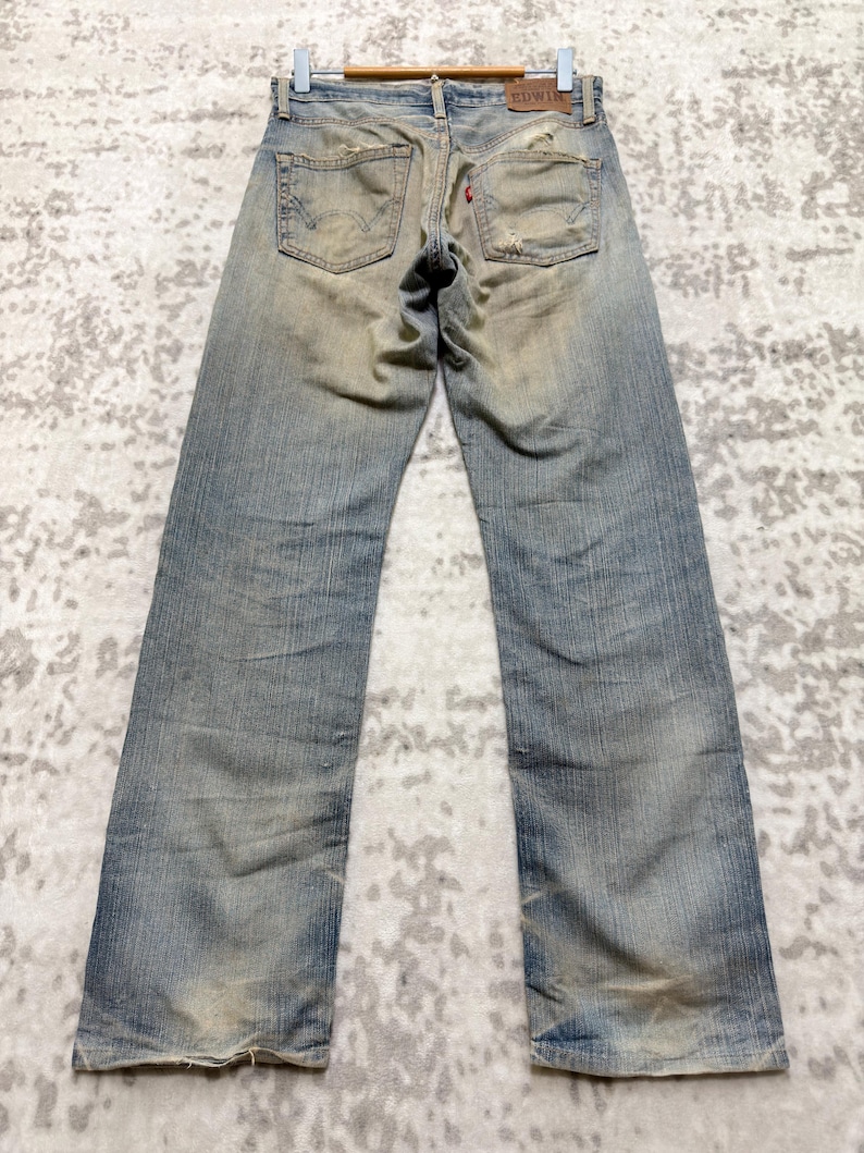 Puede incluir: Vaqueros de mezclilla azul descolorido con detalles desgastados, incluyendo parches desgastados y una etiqueta roja. Los vaqueros se muestran en una percha de madera, mostrando los bolsillos traseros y el estilo vintage. Corte recto.