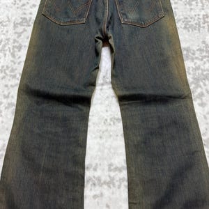 May include: Dark wash denim jeans with a brown leather Wrangler patch on the back pocket. The jeans have a classic five-pocket design and a straight leg fit. The stitching is a contrasting brown color.