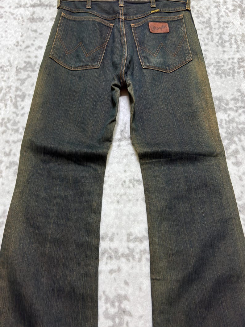 May include: Dark wash denim jeans with a brown leather Wrangler patch on the back pocket. The jeans have a classic five-pocket design and a straight leg fit. The stitching is a contrasting brown color.