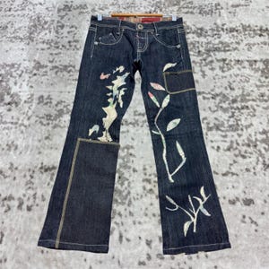 Könnte beinhalten: Dunkelblaue Jeans mit floralen Stickereien in Weiß, Rosa und Grün. Die Jeans hat ein ausgestelltes Bein, eine kleine Tasche am rechten Bein und einen kontrastierenden Aufnäher am linken Bein. Die Jeans hängt an einem Holzbügel.