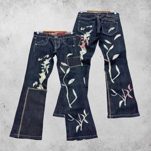 Könnte beinhalten: Dunkelblaue Jeans aus Denim mit ausgestelltem Bein, weißen Kontrastnähten und dekorativen Aufnähern mit gestickten Blumen- und Blattmustern in Rosa, Grün und Weiß. Die Jeans ist mit Vorder- und Gesäßtaschen ausgestattet.