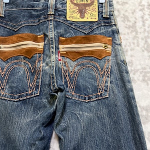 May include: Blue denim jeans with brown suede-like accents on the back pockets, featuring zippers and decorative stitching. The jeans have a faded wash and a leather patch with the brand name "EDWIN".