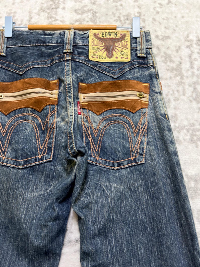 May include: Blue denim jeans with brown suede-like accents on the back pockets, featuring zippers and decorative stitching. The jeans have a faded wash and a leather patch with the brand name "EDWIN".