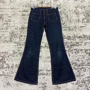 Könnte beinhalten: Dunkelblaue Schlaghose aus Denim mit zwei Vordertaschen und zwei Gesäßtaschen. Die Jeans hängt an einem Holzbügel. Der Denim hat vorne eine leichte Ausbleichung. Die Jeans hat einen Knopf- und Reißverschluss.