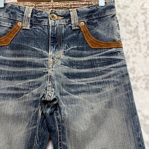 May include: Blue denim jeans with brown suede-like pocket accents and decorative stitching. The jeans have a button closure and a patterned waistband. The denim has a faded, worn appearance.