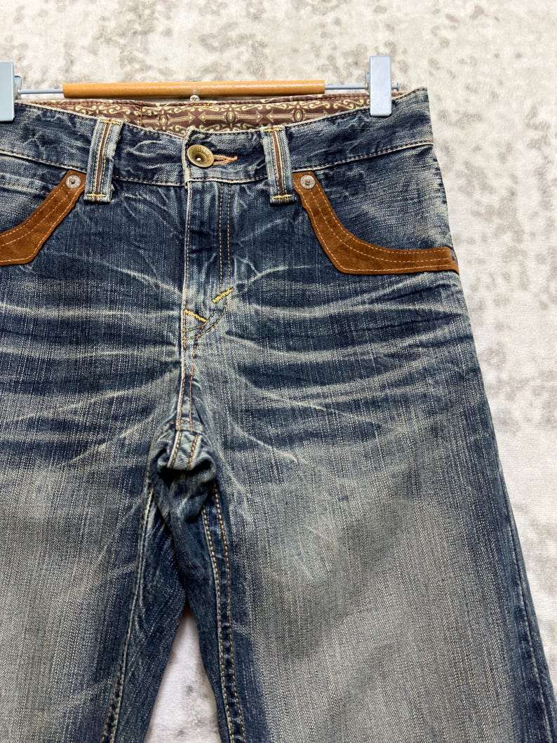 May include: Blue denim jeans with brown suede-like pocket accents and decorative stitching. The jeans have a button closure and a patterned waistband. The denim has a faded, worn appearance.