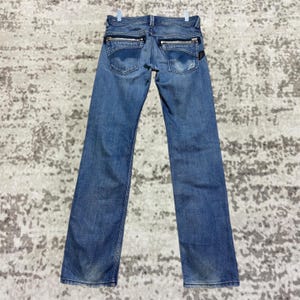 Könnte beinhalten: Blaue Jeans aus Denim mit Reißverschlusstaschen auf der Rückseite. Die Jeans haben eine mittelblaue Waschung mit leichtem Ausbleichen und hängen an einem weißen Kleiderbügel. Die Jeans haben einen geraden Beinverlauf.