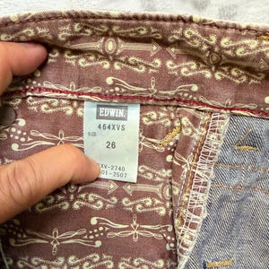 May include: Close-up of the inside of denim jeans. The jeans have a patterned lining with arrows and decorative elements. The tag reads "EDWIN 464XVS SIZE 26". The denim is a faded blue color.