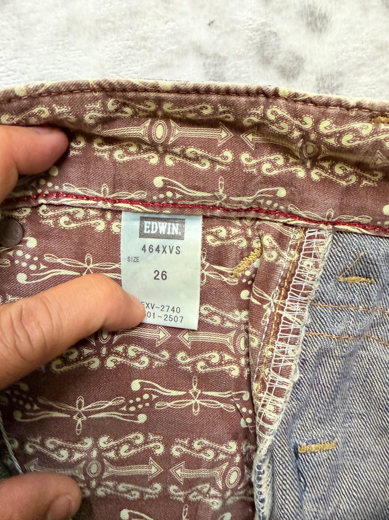 May include: Close-up of the inside of denim jeans. The jeans have a patterned lining with arrows and decorative elements. The tag reads "EDWIN 464XVS SIZE 26". The denim is a faded blue color.