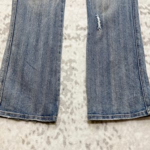 May include: Light blue denim jeans with a slight flare at the bottom. The jeans have a distressed area on the right leg. The jeans are laid flat on a patterned surface.