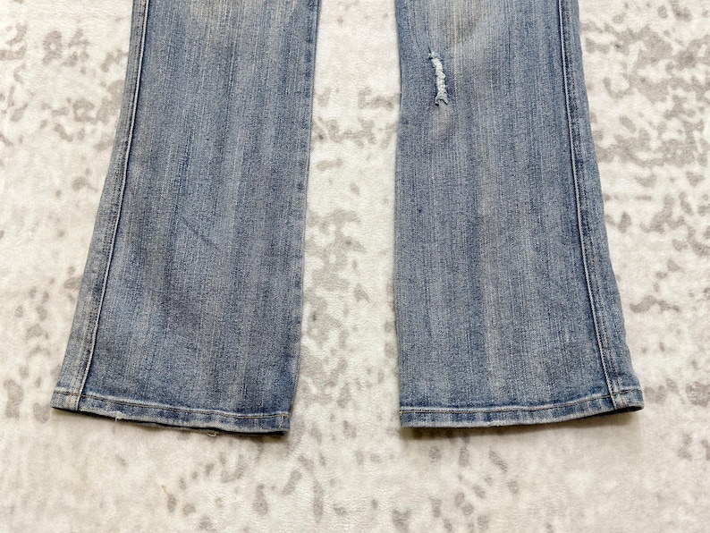 May include: Light blue denim jeans with a slight flare at the bottom. The jeans have a distressed area on the right leg. The jeans are laid flat on a patterned surface.