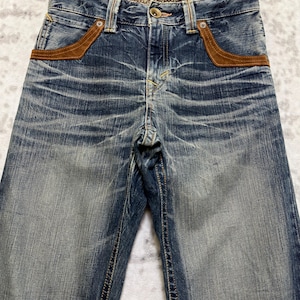 May include: Faded blue denim jeans with brown suede-like pocket accents. The jeans have a button closure and a decorative patterned waistband. The denim has a distressed look with visible wear and tear, giving them a vintage aesthetic.
