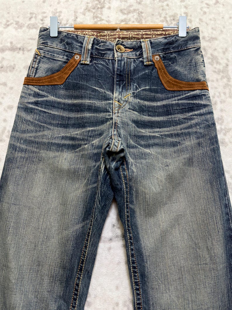 May include: Faded blue denim jeans with brown suede-like pocket accents. The jeans have a button closure and a decorative patterned waistband. The denim has a distressed look with visible wear and tear, giving them a vintage aesthetic.