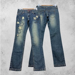 Könnte beinhalten: Blaue Jeans mit verwaschener Optik und ausgestelltem Bein. Die Jeans haben sternförmige Aufnäher in verschiedenen Größen auf der Vorderseite. Auf der Rückseite der Jeans befindet sich ein kleiner Sternaufnäher auf der Gesäßtasche. Die Jeans hängen an einem Holzbügel.