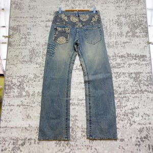 Könnte beinhalten: Hellblaue Jeans mit dekorativer Stickerei auf den Gesäßtaschen und am Bund. Die Jeans hat einen geraden Beinverlauf und eine verwaschene Waschung. Auf der linken Tasche befindet sich ein Aufnäher. Die Jeans hängt an einem Holzbügel.