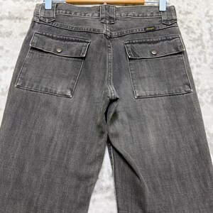 May include: A pair of gray denim jeans with two square pockets on the back, each secured with a single button. The jeans are hanging on a wooden hanger against a light-colored background.