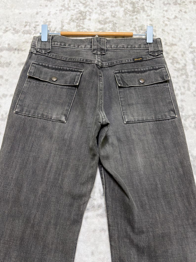 May include: A pair of gray denim jeans with two square pockets on the back, each secured with a single button. The jeans are hanging on a wooden hanger against a light-colored background.