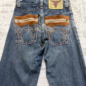 May include: Blue denim jeans with brown suede-like accents on the back pockets. The jeans feature a zipper detail on the pockets and a leather patch with the brand name "Edwin". The stitching is a contrasting orange color.