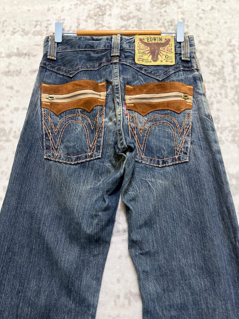 May include: Blue denim jeans with brown suede-like accents on the back pockets. The jeans feature a zipper detail on the pockets and a leather patch with the brand name "Edwin". The stitching is a contrasting orange color.