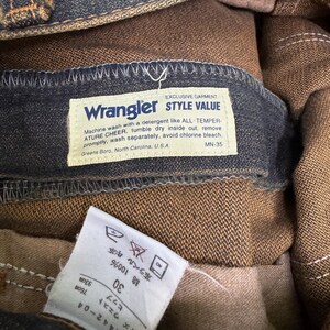 May include: Close-up of a pair of Wrangler jeans. The label reads "Wrangler STYLE VALUE" and includes washing instructions. The jeans are a dark blue denim with brown interior lining. The size tag indicates a waist size of 30 inches.