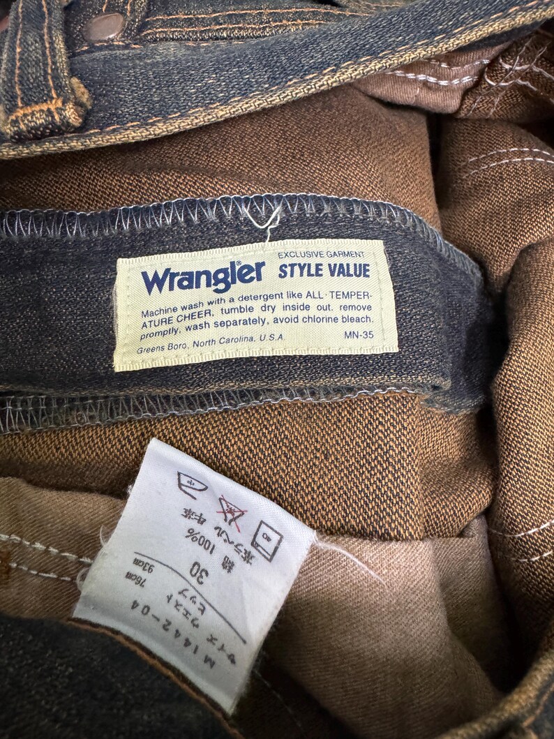 May include: Close-up of a pair of Wrangler jeans. The label reads "Wrangler STYLE VALUE" and includes washing instructions. The jeans are a dark blue denim with brown interior lining. The size tag indicates a waist size of 30 inches.