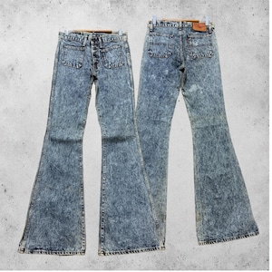 Könnte beinhalten: Eine Jeans aus blauem Acid-Wash-Denim mit hoher Taille und ausgestelltem Bein. Die Vorderseite zeigt zwei aufgesetzte Taschen und einen Knopfverschluss, während die Rückseite zwei Gesäßtaschen und ein braunes Lederetikett am Bund aufweist.