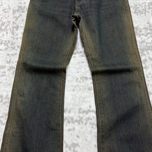 May include: Dark wash denim jeans with a slight flare. The jeans have a classic five-pocket design and a button and zipper closure. The denim has a faded, worn look with subtle brown accents along the seams and edges.