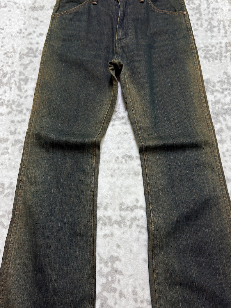 May include: Dark wash denim jeans with a slight flare. The jeans have a classic five-pocket design and a button and zipper closure. The denim has a faded, worn look with subtle brown accents along the seams and edges.