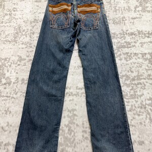 May include: Blue denim jeans with brown suede-like accents on the back pockets and waistband. The jeans have a classic five-pocket design and a straight leg fit. A leather patch with text is visible on the back.