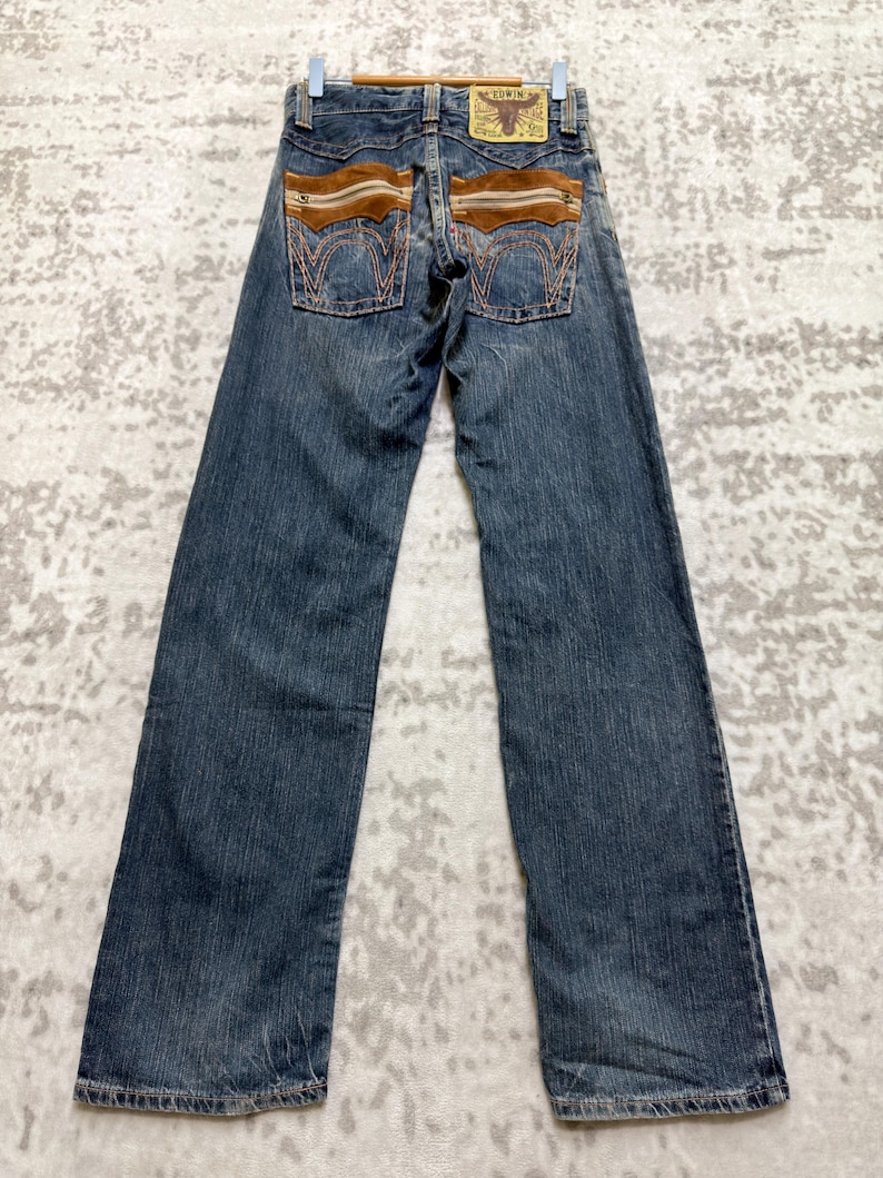 May include: Blue denim jeans with brown suede-like accents on the back pockets and waistband. The jeans have a classic five-pocket design and a straight leg fit. A leather patch with text is visible on the back.