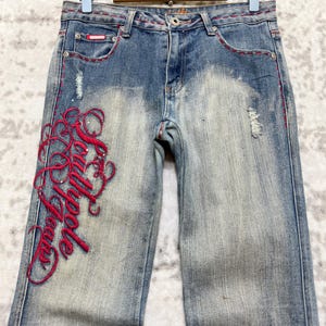 May include: Faded blue denim jeans with red embroidered script on the left leg. The jeans have a distressed look with some fading and small tears. Red stitching accents the pockets and seams. The jeans are hanging on a wooden hanger.