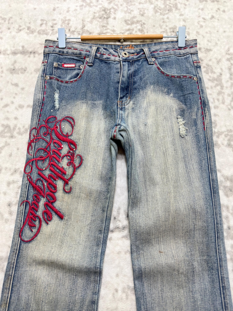 May include: Faded blue denim jeans with red embroidered script on the left leg. The jeans have a distressed look with some fading and small tears. Red stitching accents the pockets and seams. The jeans are hanging on a wooden hanger.