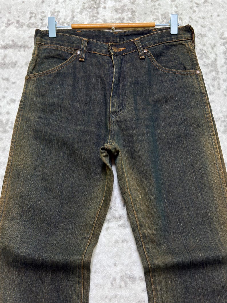 May include: Dark wash denim jeans with a faded, worn appearance. The jeans have a classic five-pocket design, a button and zipper closure, and belt loops. The denim has a slight sheen, and the jeans are hanging on a hanger.