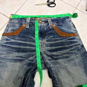 May include: Blue denim jeans with brown suede-like pocket accents. A green measuring tape shows the waist size. Scissors and a box are in the background. The jeans are distressed and have a button and zipper closure.