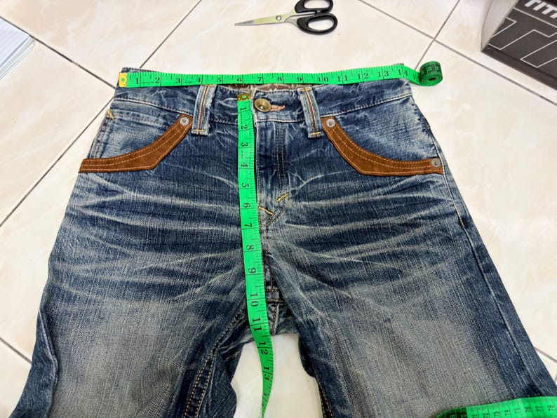 May include: Blue denim jeans with brown suede-like pocket accents. A green measuring tape shows the waist size. Scissors and a box are in the background. The jeans are distressed and have a button and zipper closure.