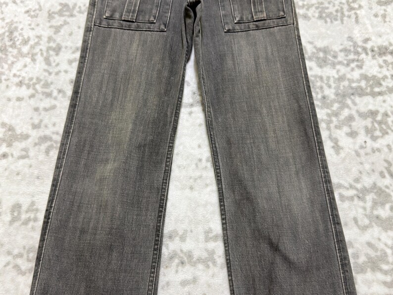 May include: A pair of gray denim jeans with two square pockets on the front. The jeans have a straight leg cut and a faded appearance. The stitching is visible along the seams and pockets.