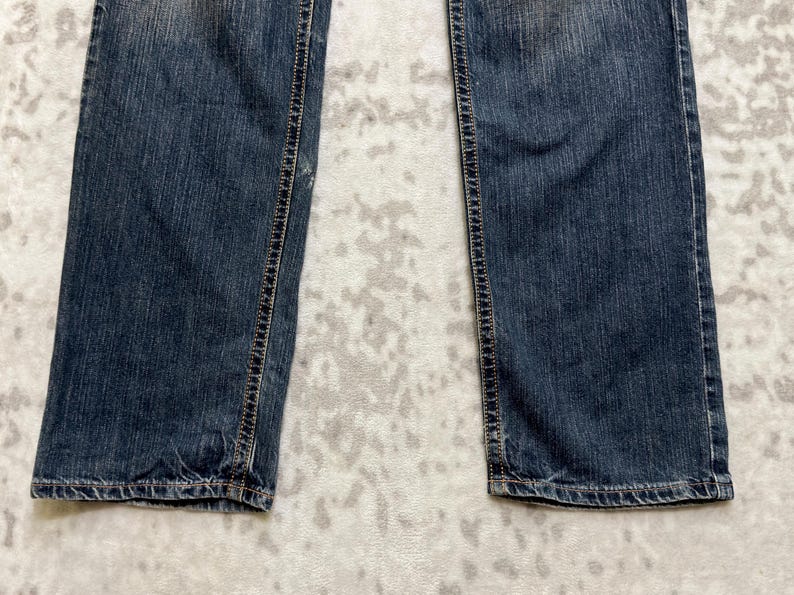 May include: Close-up of the lower portion of a pair of blue denim jeans. The jeans have a classic five-pocket design with visible stitching along the seams and a faded wash. The fabric appears worn and soft.