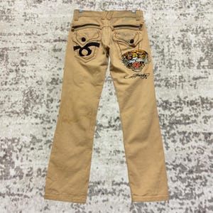 Könnte beinhalten: Beige Denim-Jeans mit geradem Bein. Die Gesäßtaschen haben schwarze Stickereien, darunter ein Tigerkopf und der Markenname. Die Jeans hat Reißverschlusstaschen und dekorative Nähte.