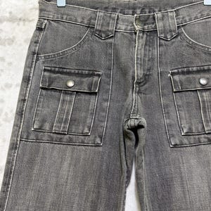 May include: Close-up of gray denim pants with two front pockets and two cargo pockets. The pants have a button closure and belt loops. The stitching is white, and the pants are hanging on a hanger.