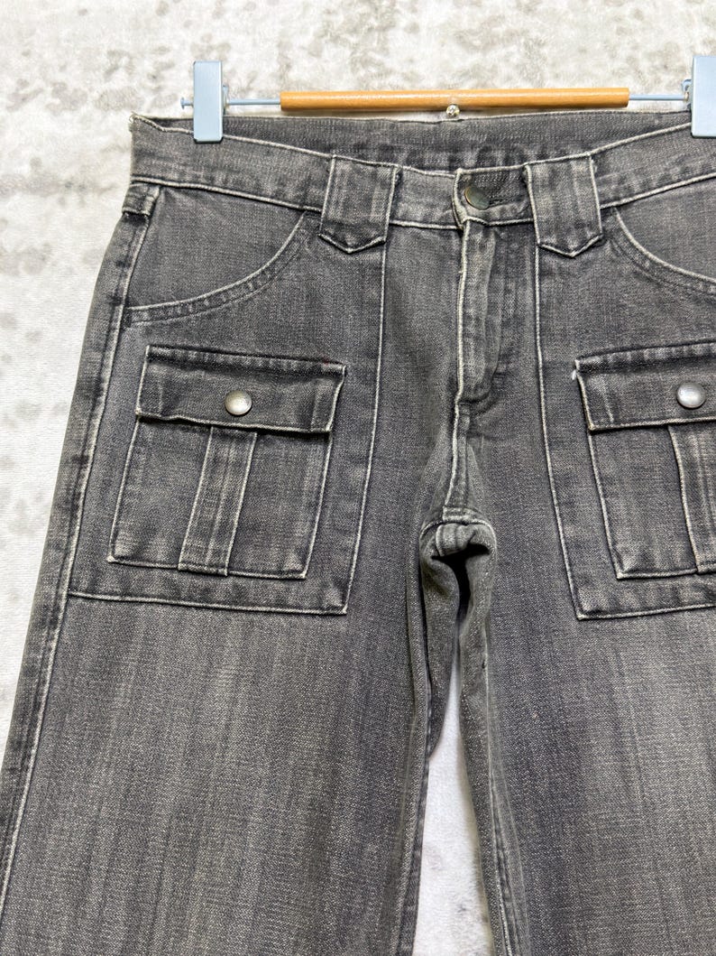 May include: Close-up of gray denim pants with two front pockets and two cargo pockets. The pants have a button closure and belt loops. The stitching is white, and the pants are hanging on a hanger.