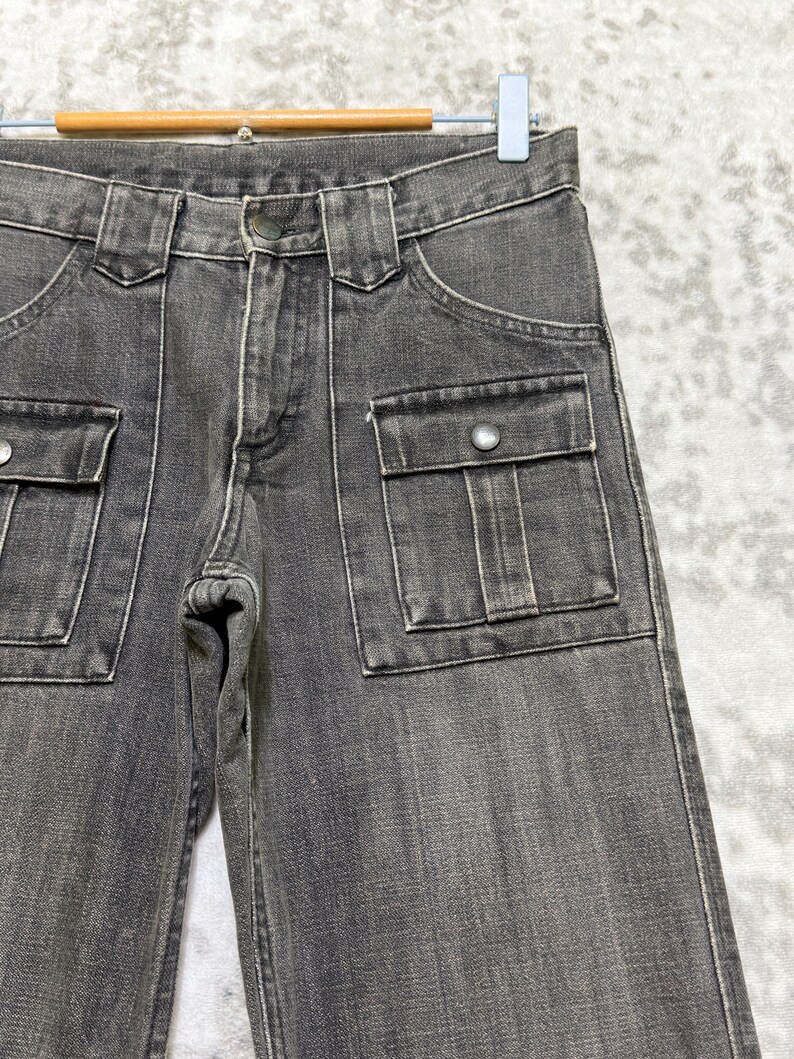 May include: Close-up of gray denim pants with two front pockets and two side pockets. The pants have a button closure and belt loops. The denim is a faded gray color.