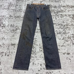 Puede incluir: Vaqueros de mezclilla de lavado oscuro con bragueta de botones, diseño clásico de cinco bolsillos y pernera recta. Los vaqueros se muestran en una percha de madera, mostrando toda la longitud y los detalles de la prenda. La mezclilla tiene un aspecto ligeramente desgastado.