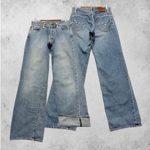 May include: Two pairs of light blue denim jeans hanging on wooden hangers. One pair has a button fly and a wide leg. The other pair has a classic five-pocket design with a straight leg. Both pairs show signs of wear.