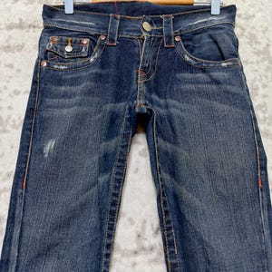 May include: Dark wash denim jeans with orange stitching and metal rivets. The jeans have a classic five-pocket design and are displayed on a hanger. The denim has a distressed look with some fading and wear.