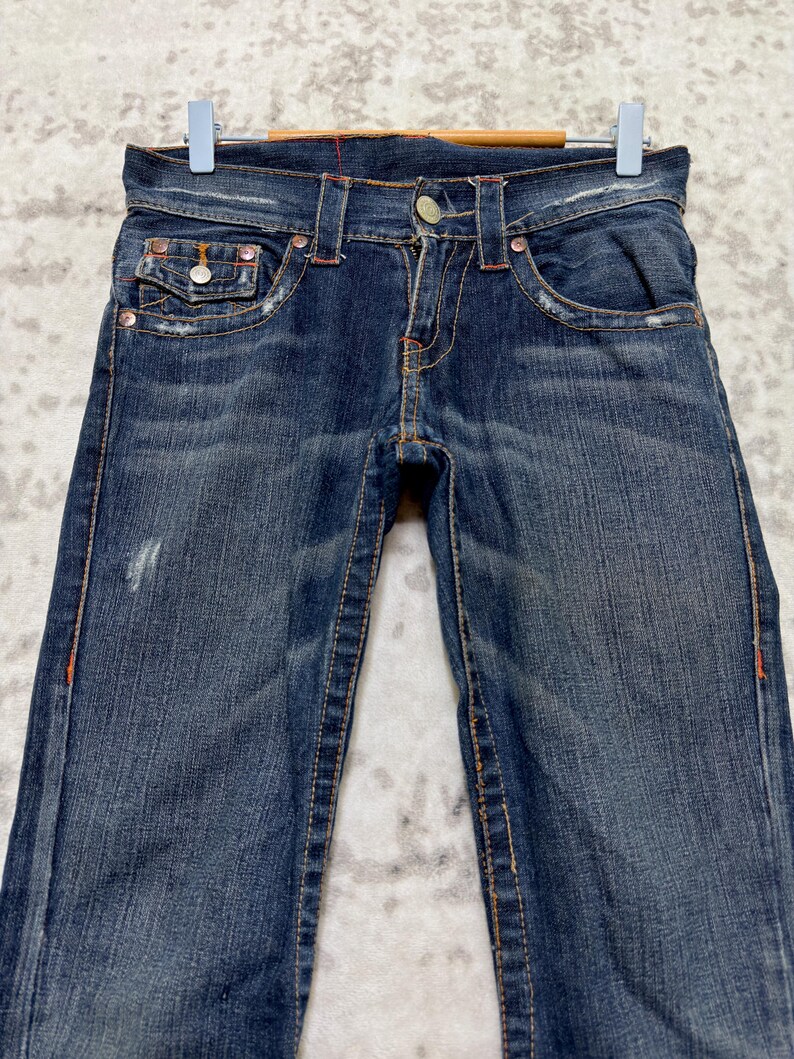 May include: Dark wash denim jeans with orange stitching and metal rivets. The jeans have a classic five-pocket design and are displayed on a hanger. The denim has a distressed look with some fading and wear.
