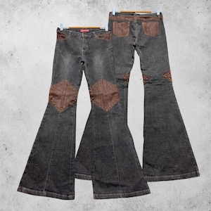 Könnte beinhalten: Zwei dunkelgraue Schlaghosen aus Denim mit braunen, lederartigen Einsätzen an den vorderen Oberschenkeln und Gesäßtaschen. Die vorderen Einsätze sind rautenförmig mit Metallnieten verziert, während die Gesäßtaschen rechteckig geformt sind.